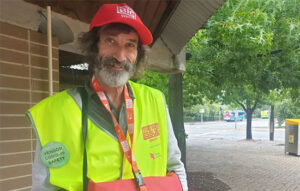 A vendor wearing his Big Issue uniform - a red cap and fluorescent yellow vest both with The Big Issue logo.